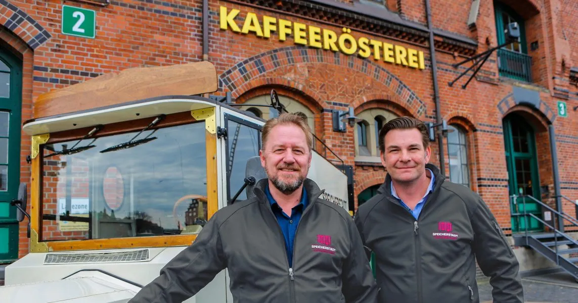 Zwei Männer stehen vor einem Gebäude mit der Aufschrift „KAFFEERÖSTEREI“.