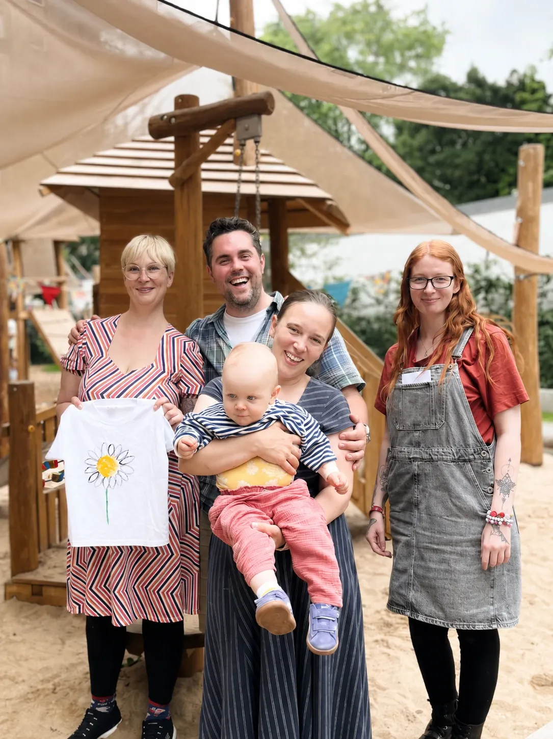 Eine Gruppe von vier Erwachsenen und einem Baby posiert lächelnd auf einem Spielplatz.