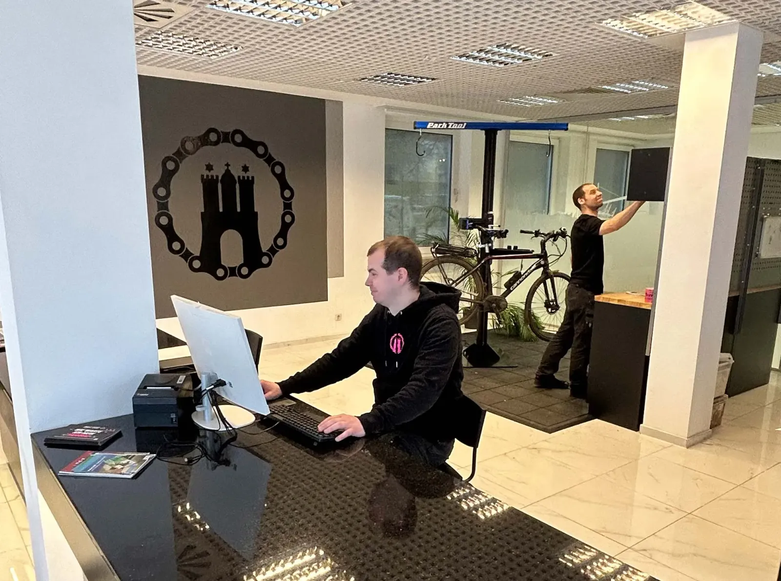Two employees work in the workshop, one at the computer and the other on a bicycle.